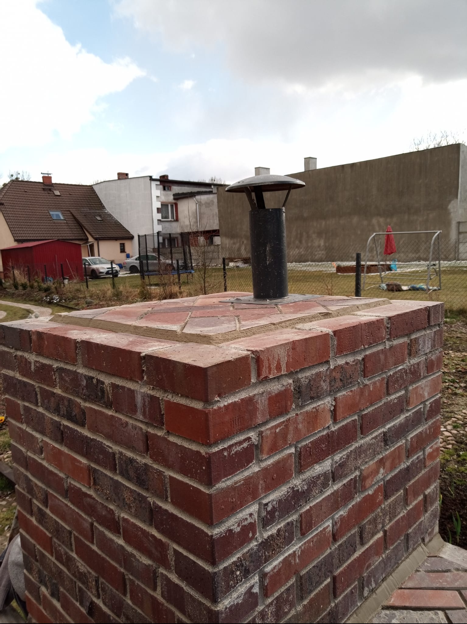 Murowany komin z czerwonej cegły z czarnym wkładem kominowym i daszkiem na zewnątrz, na tle zabudowań i pochmurnego nieba.
