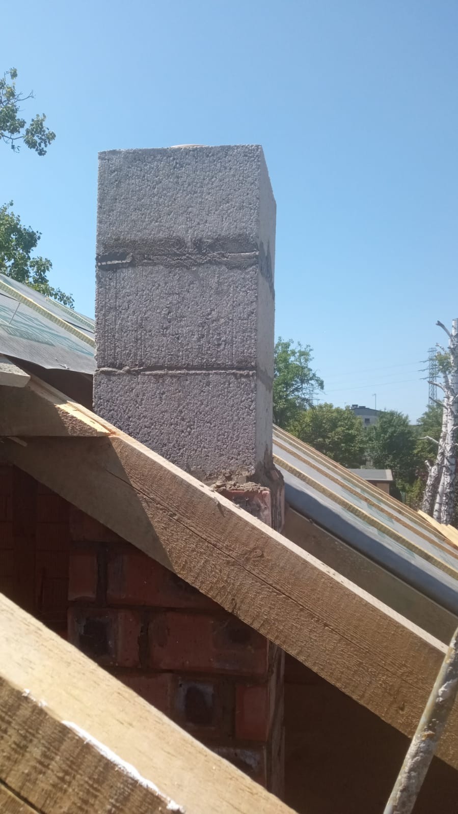 Wznoszenie komina z bloczków betonowych na dachu z drewnianą konstrukcją. Widoczne elementy izolacji i ceglana podstawa komina na tle błękitnego nieba.