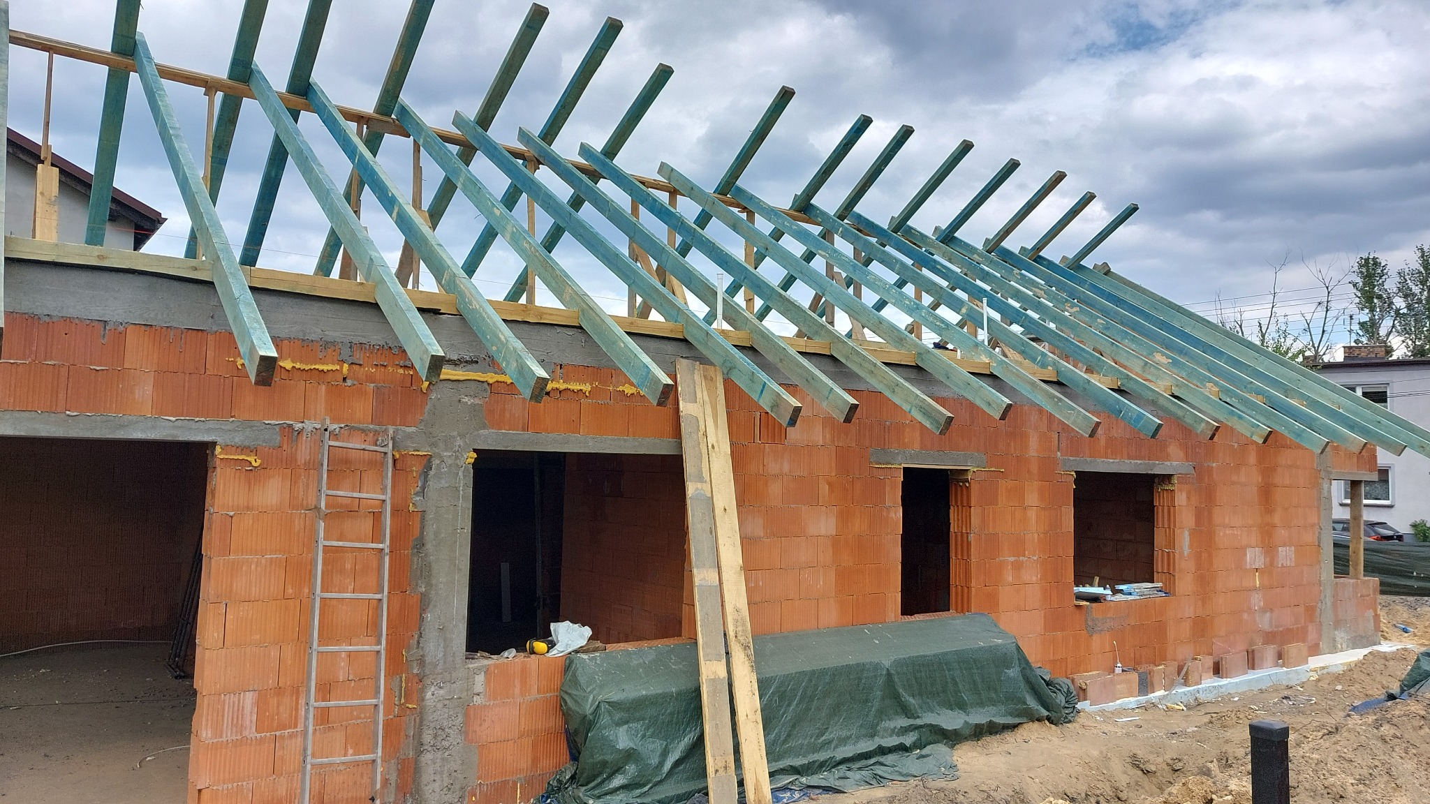 Ceglany budynek w stanie surowym otwartym z drewnianą konstrukcją dachu, drabina oparta o ścianę, widok na budowę w słoneczny dzień.