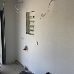 Energofuse Technology - Rozdzielnia elektryczna w nowym budynku, widok z boku, z otwartymi drzwiami garażowymi i puszkami instalacyjnymi. W tle zieleń i błękit nieba.