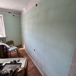 Energofuse Technology - Ściana z otworami pod instalację elektryczną w pokoju z drewnianą podłogą i meblami. Widoczne narzędzia i kable. Przygotowanie do montażu gniazdek.