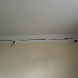 Energofuse Technology - Fragment ściany z widocznymi kablami elektrycznymi poprowadzonymi pod sufitem. Dwa otwory w ścianie z wystającymi przewodami.