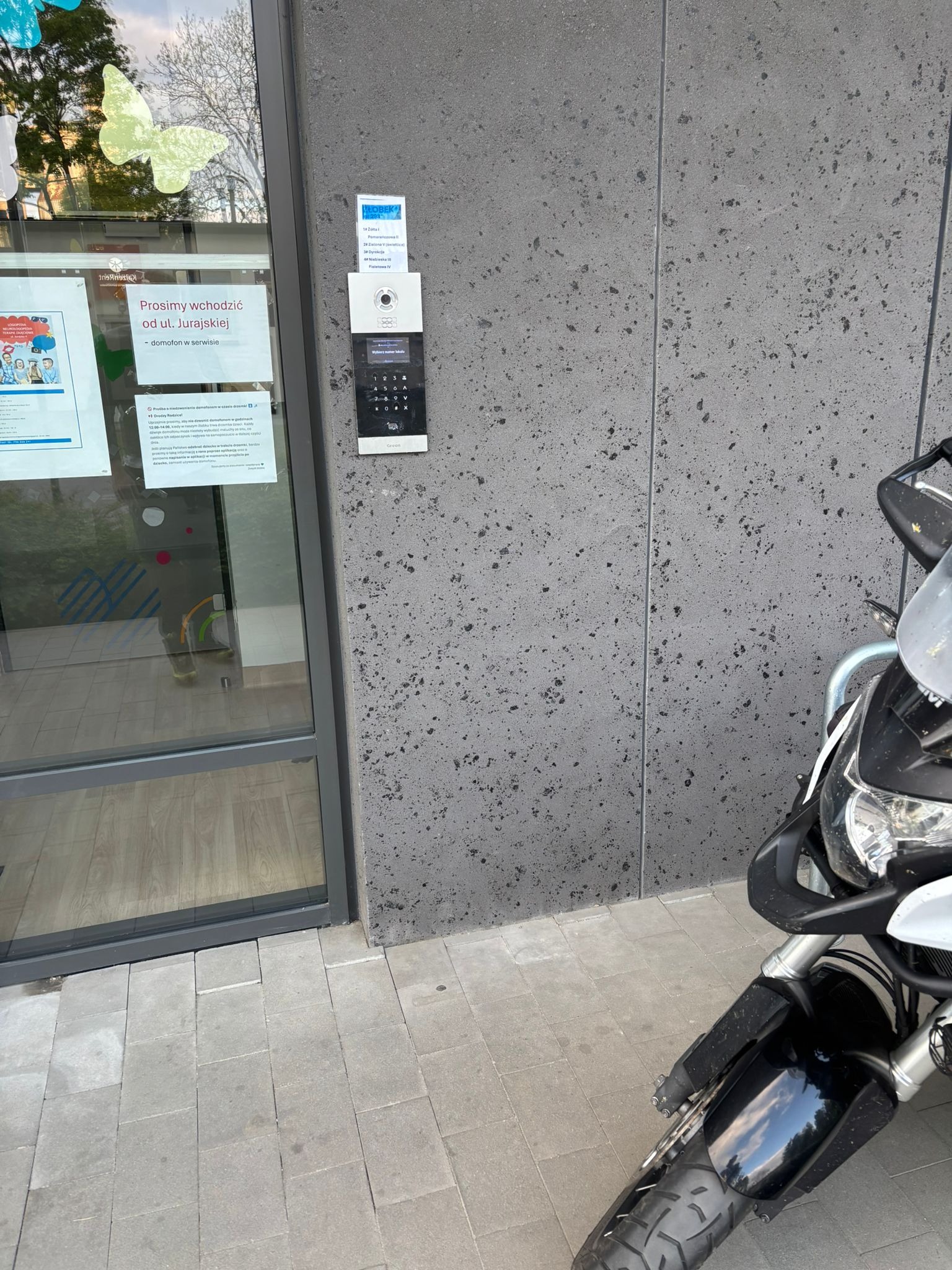 Domofon z klawiaturą numeryczną na szarej ścianie budynku, obok drzwi wejściowych z informacją o serwisie. Część motocykla widoczna z prawej strony.