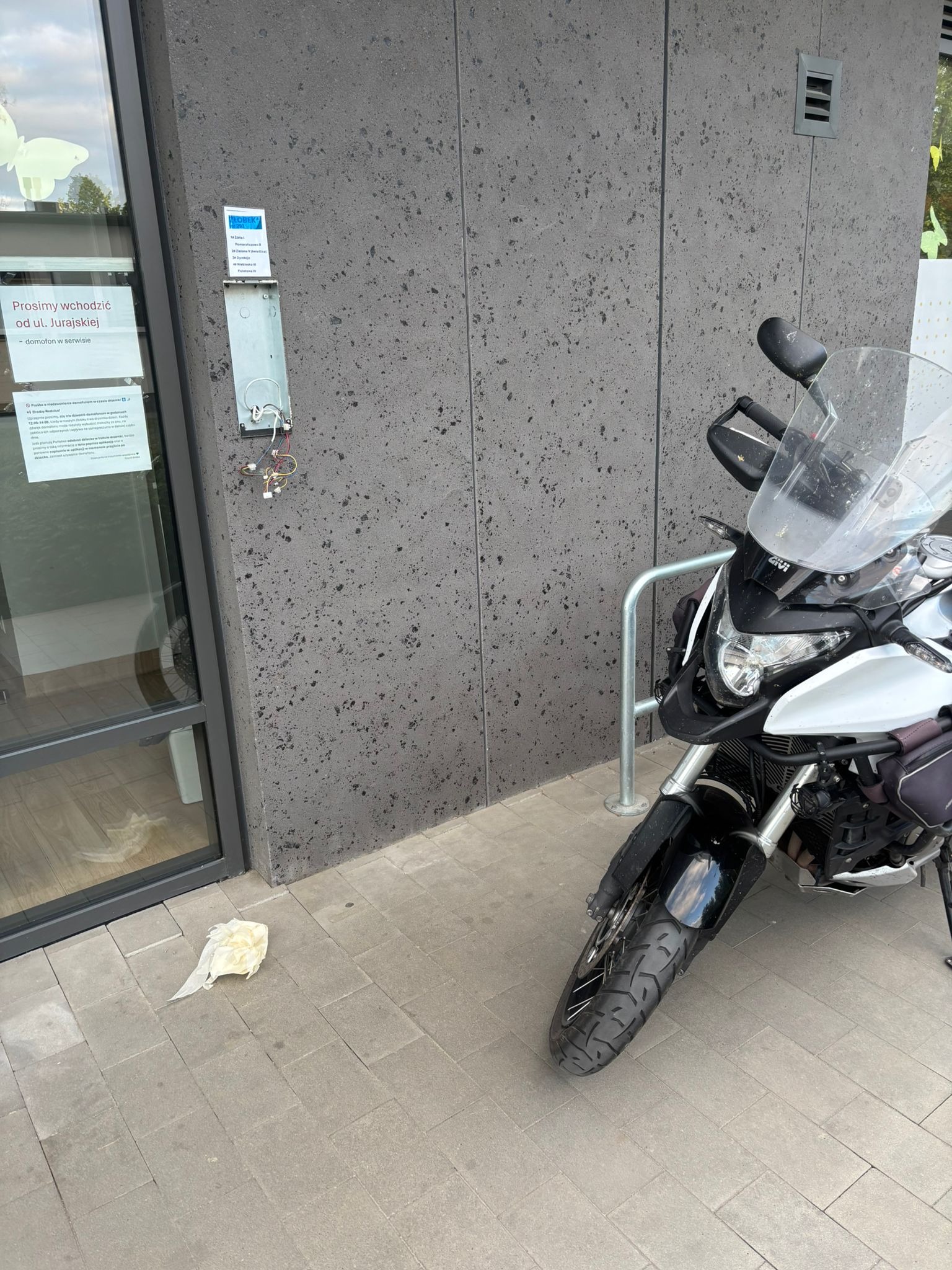 Uszkodzony domofon z wystającymi kablami na szarej ścianie budynku, obok zaparkowany motocykl. Na drzwiach kartka z informacją.