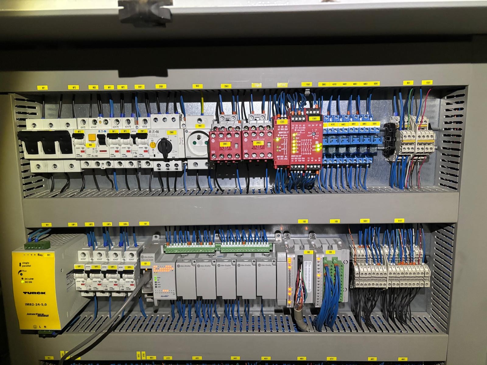Szafa sterownicza z modułami PLC Allen-Bradley, przekaźnikami bezpieczeństwa Turck i zabezpieczeniami Eaton. Widoczne liczne przewody i oznaczenia.