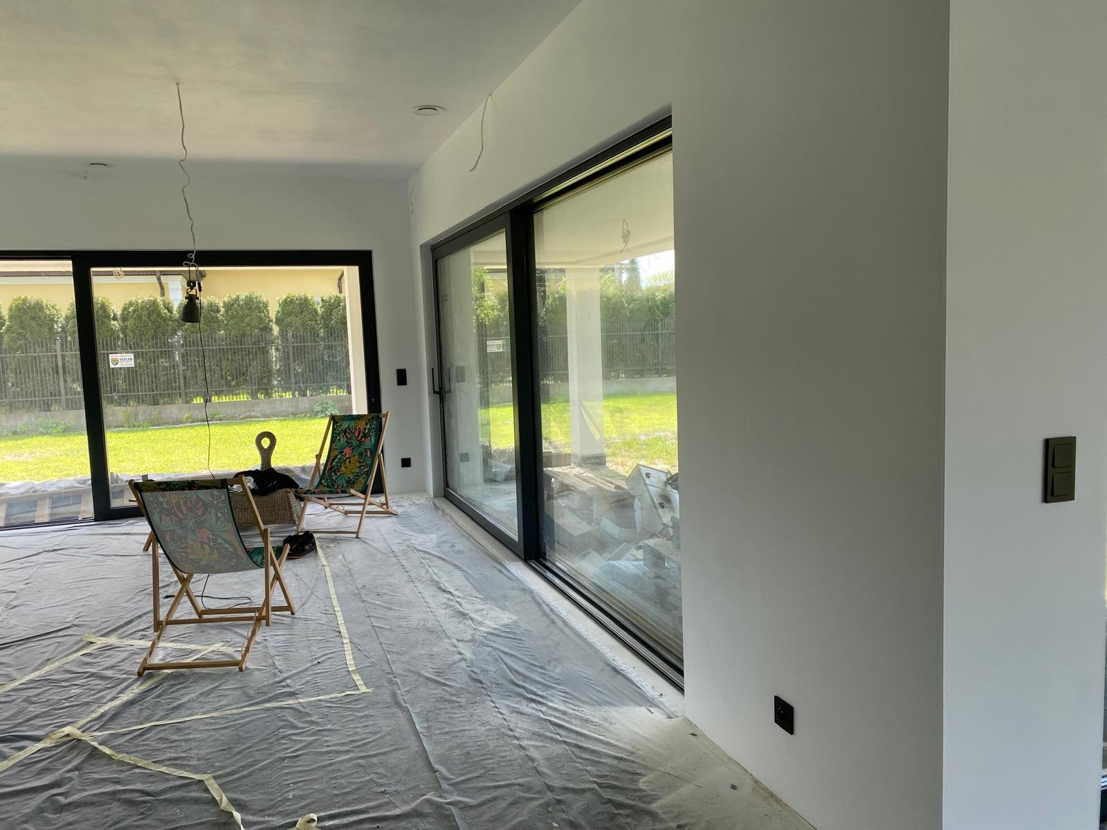 Jasne wnętrze z dużymi oknami po malowaniu, zabezpieczone folią. Widoczne krzesła plażowe i wystające kable, sugerujące prace elektryczne w toku.