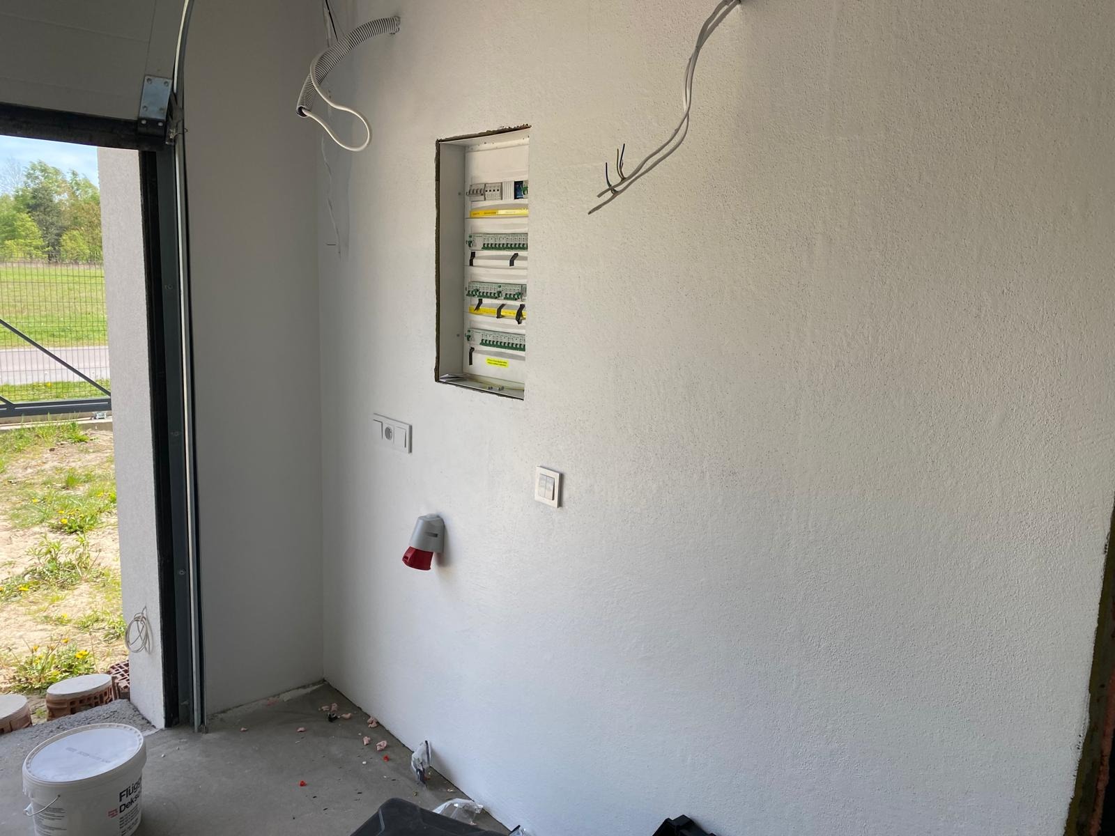 Rozdzielnia elektryczna w nowym budynku, widok z boku, z otwartymi drzwiami garażowymi i puszkami instalacyjnymi. W tle zieleń i błękit nieba.