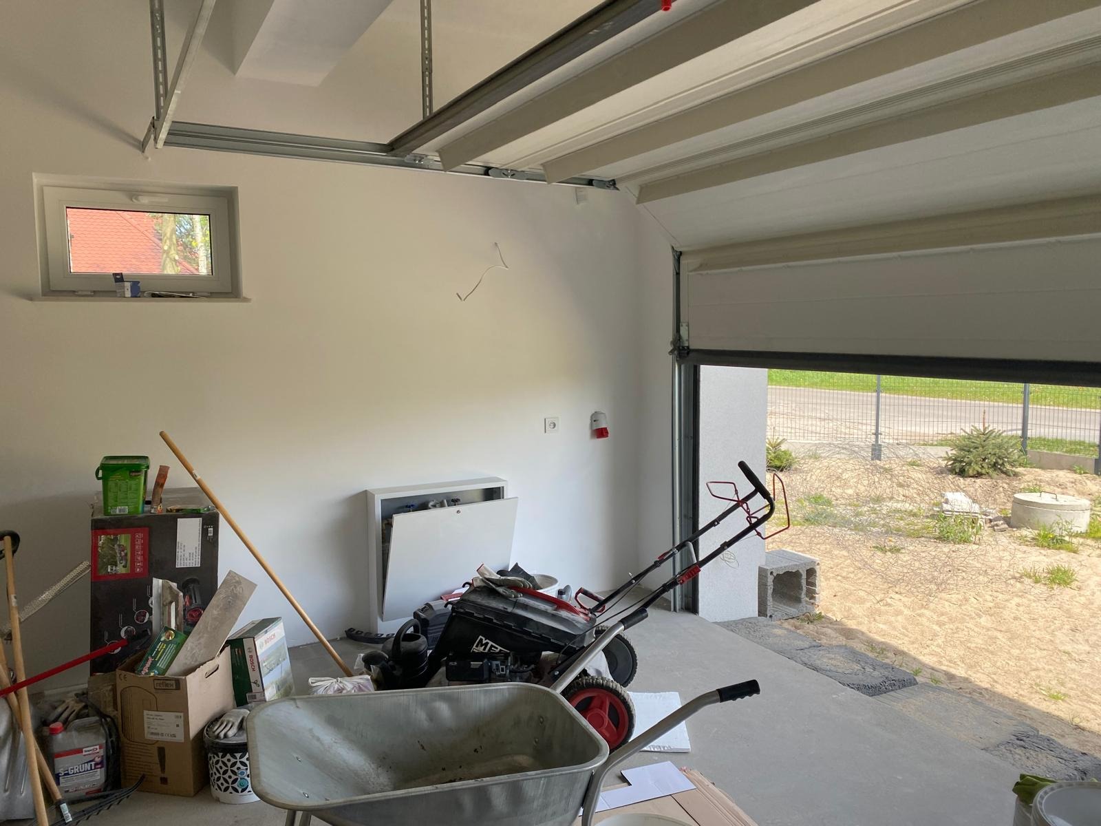 Wnętrze garażu w trakcie prac wykończeniowych: widoczna brama garażowa, instalacja elektryczna, narzędzia i materiały budowlane, sugerujące etap przygotowania do użytkowania.