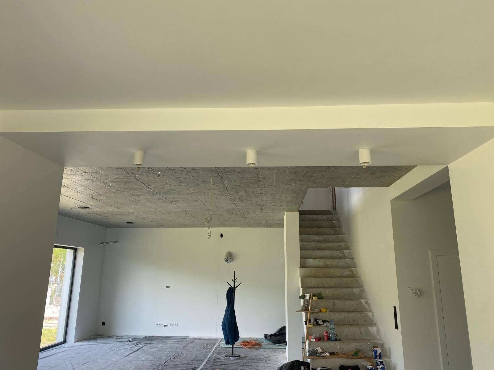 Surowy strop betonowy z widoczną instalacją elektryczną, trzy białe lampy sufitowe, nieotynkowane schody w przestronnym wnętrzu domu w trakcie wykańczania.
