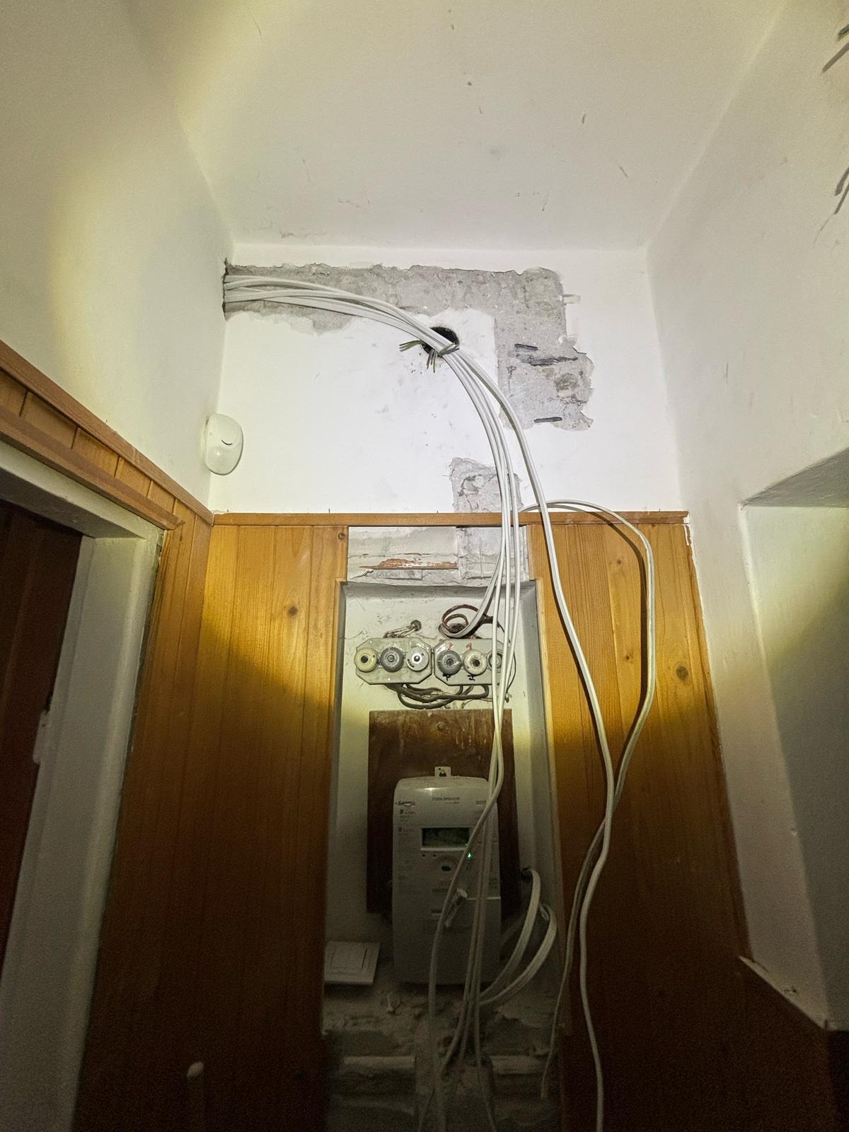 Białe kable elektryczne wychodzące ze ściany, podłączone do starej skrzynki z bezpiecznikami i licznika w drewnianej obudowie. Widoczne ślady po kuciu w ścianie.