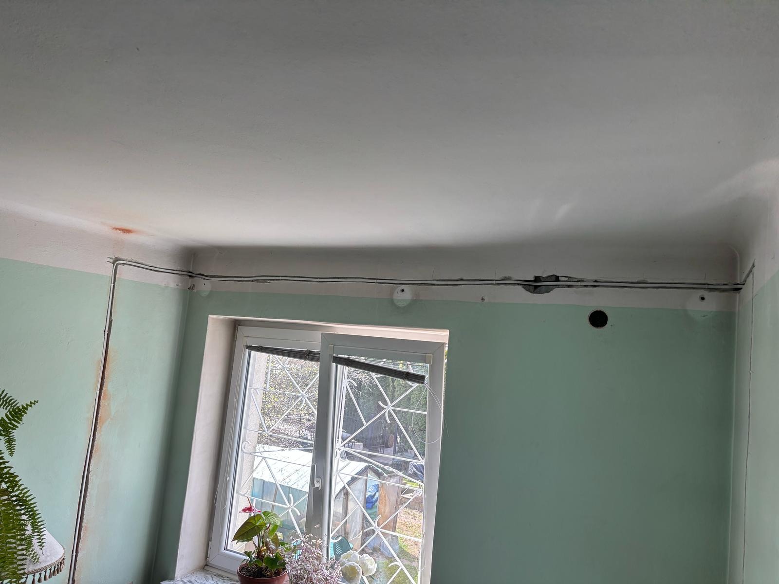 Kable elektryczne poprowadzone po ścianie wzdłuż sufitu w pokoju z oknem. Widoczne otwory w ścianie i elementy instalacji. Ściany pomalowane na zielono.