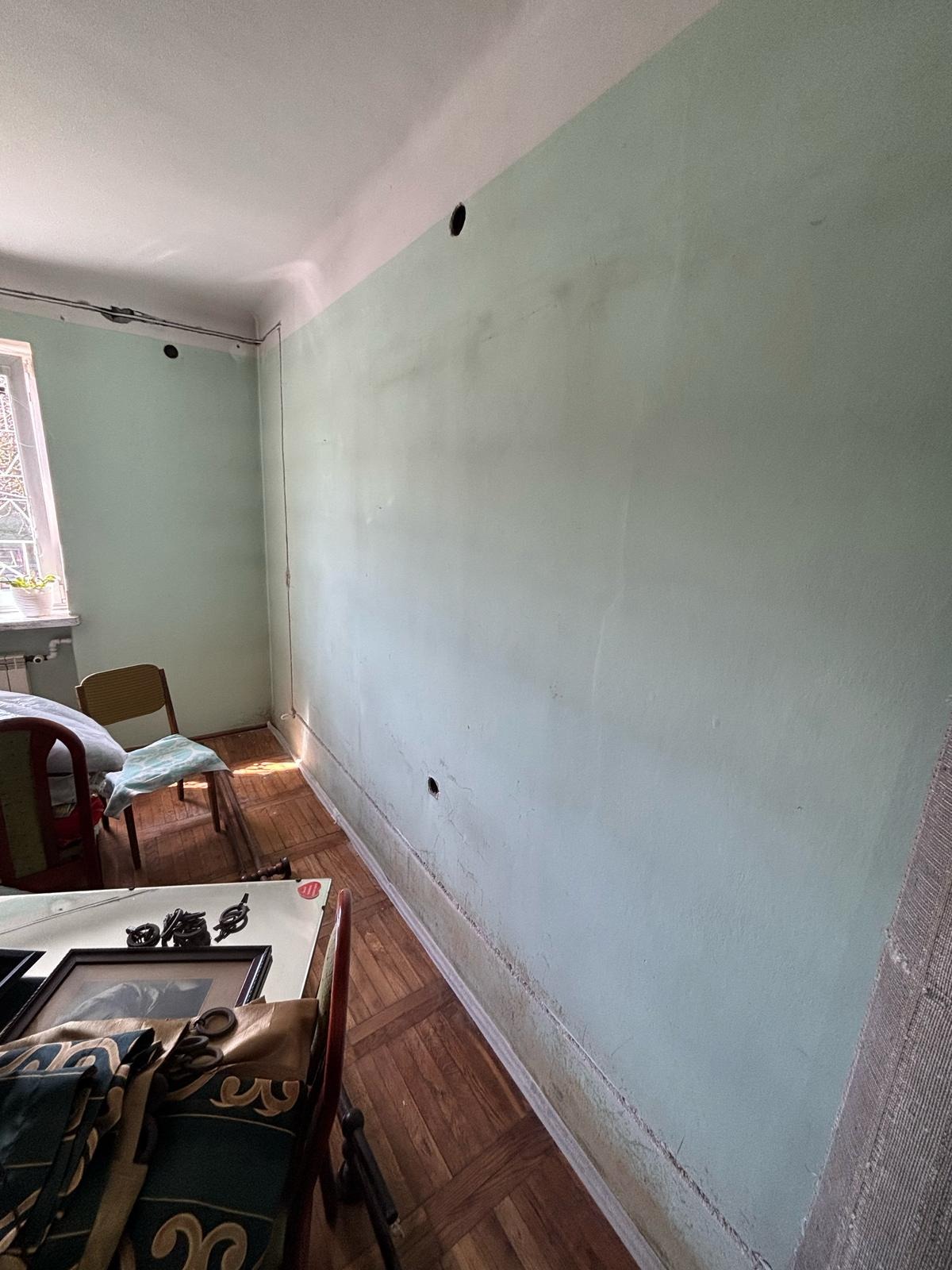 Ściana z otworami pod instalację elektryczną w pokoju z drewnianą podłogą i meblami. Widoczne narzędzia i kable. Przygotowanie do montażu gniazdek.