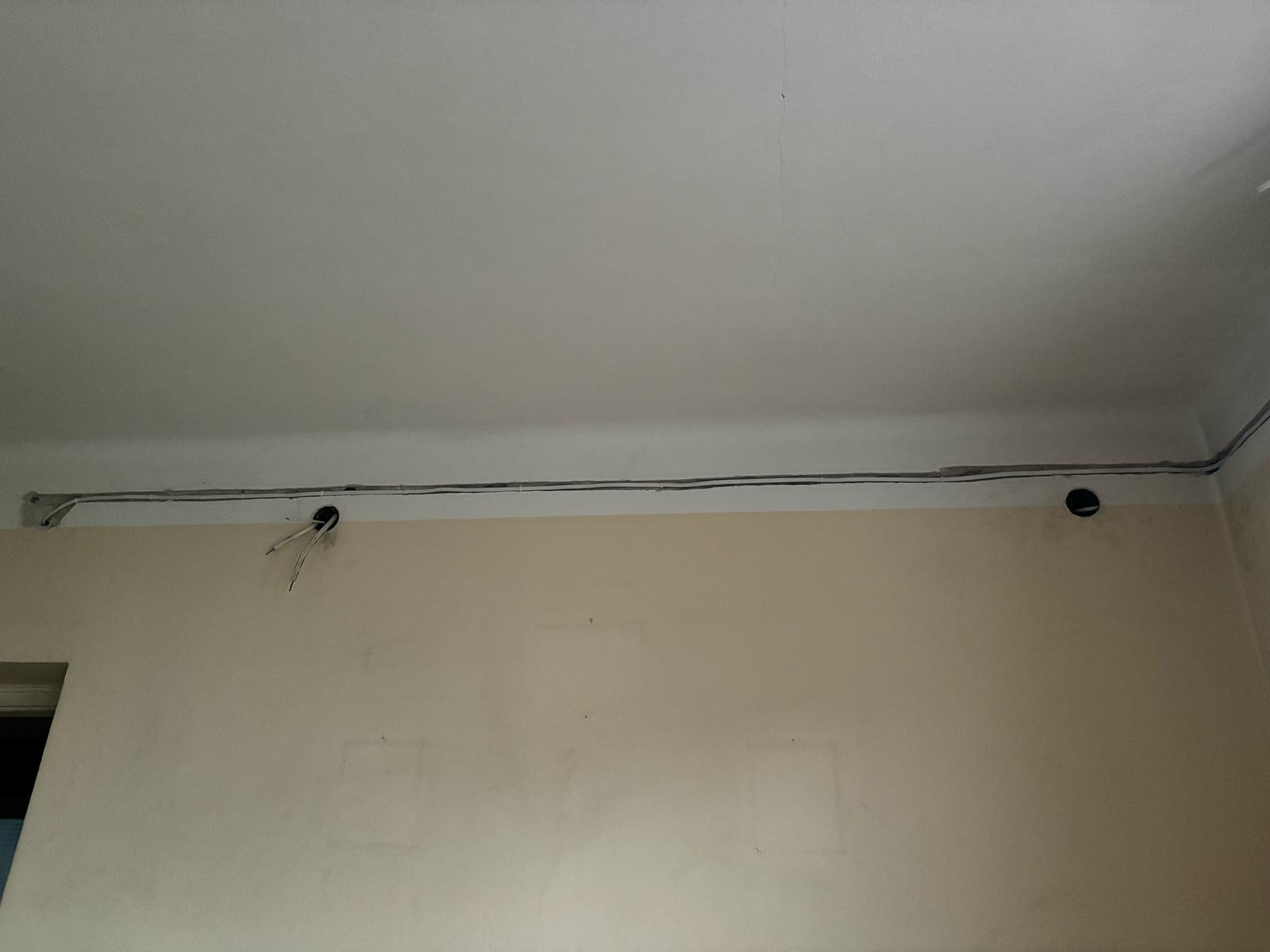 Fragment ściany z widocznymi kablami elektrycznymi poprowadzonymi pod sufitem. Dwa otwory w ścianie z wystającymi przewodami.