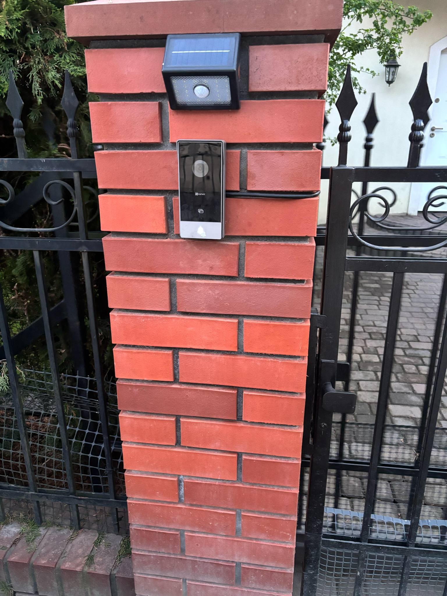 Brama z ceglanym słupkiem, na którym zamontowany jest wideodomofon z lampą solarną. Widoczne okablowanie i elementy instalacji.