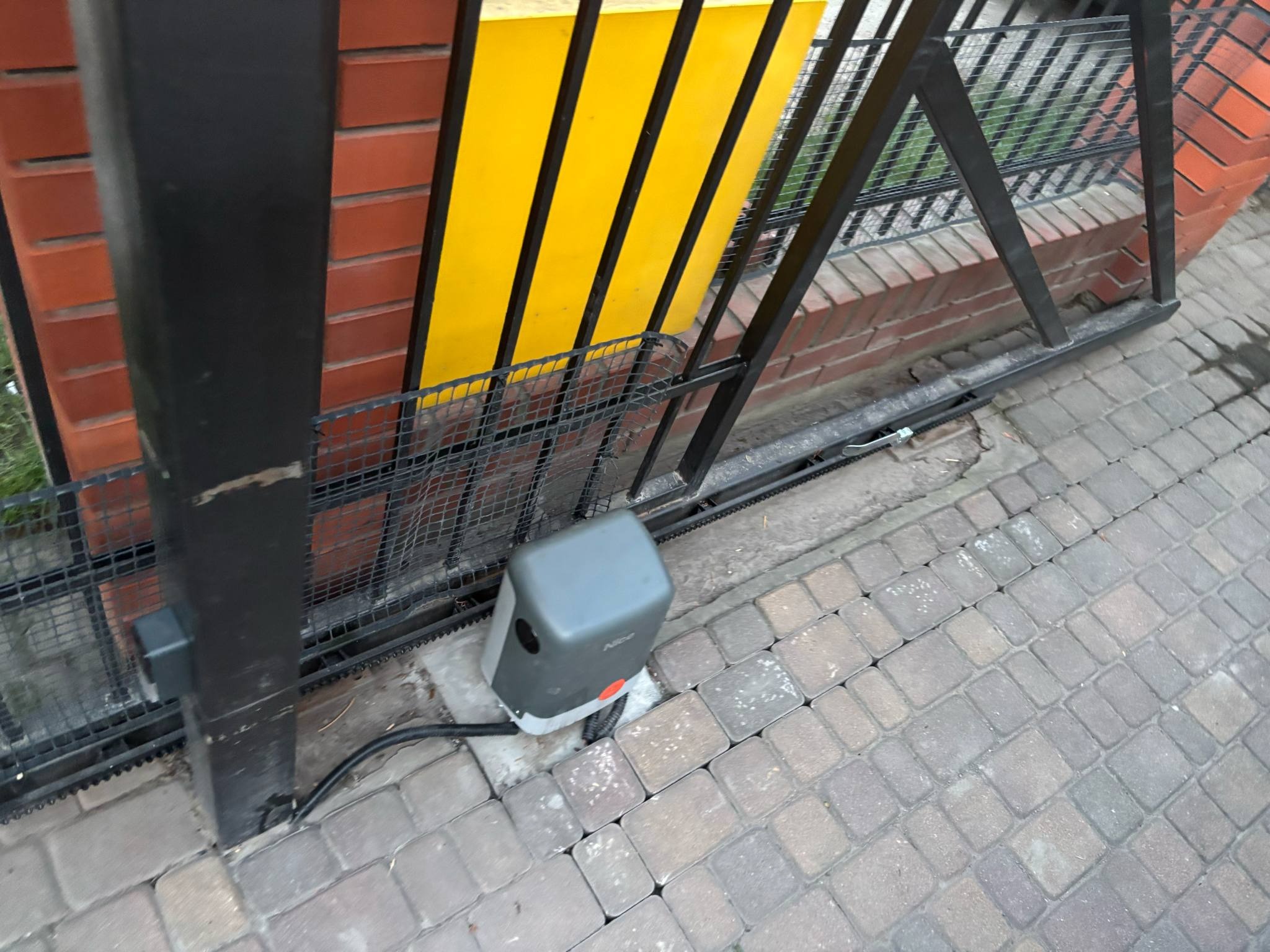 Automat do bramy przesuwnej Nice Robuskit 600 z listwą zębatą, zamontowany na kostce brukowej przy metalowej bramie z wypełnieniem siatkowym i żółtymi elementami.