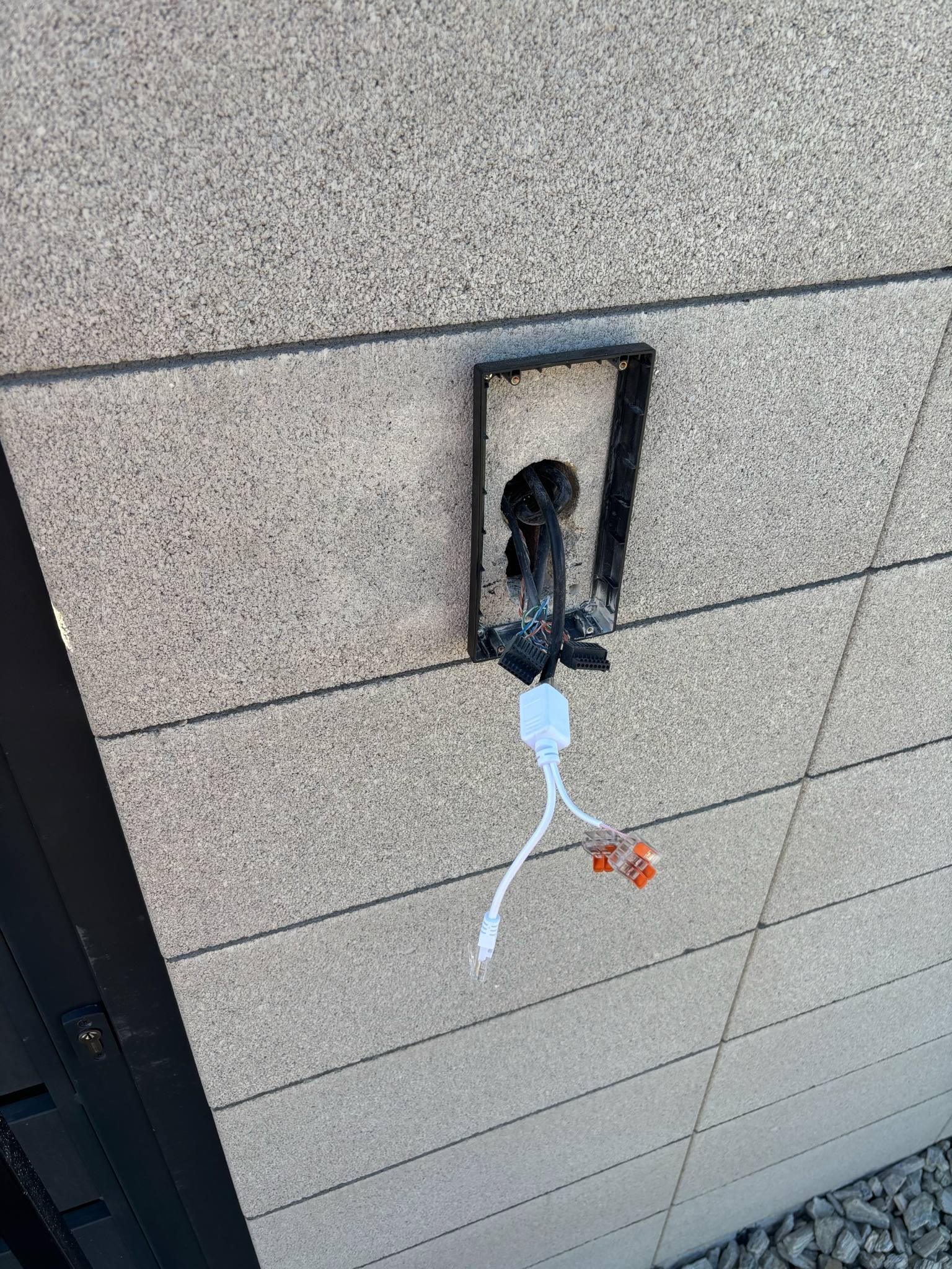 Wystająca instalacja elektryczna ze ściany z szarą elewacją. Widoczne przewody, złączki i biała kostka. Czarna ramka montażowa przytwierdzona do ściany.