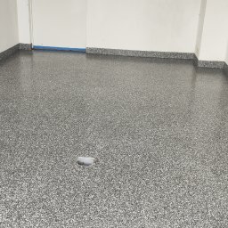 Daniel Sojko - Błyszcząca, szara posadzka z drobnym wzorem w pomieszczeniu z białymi ścianami. Widoczny odpływ podłogowy i fragment niebieskiej taśmy zabezpieczającej.