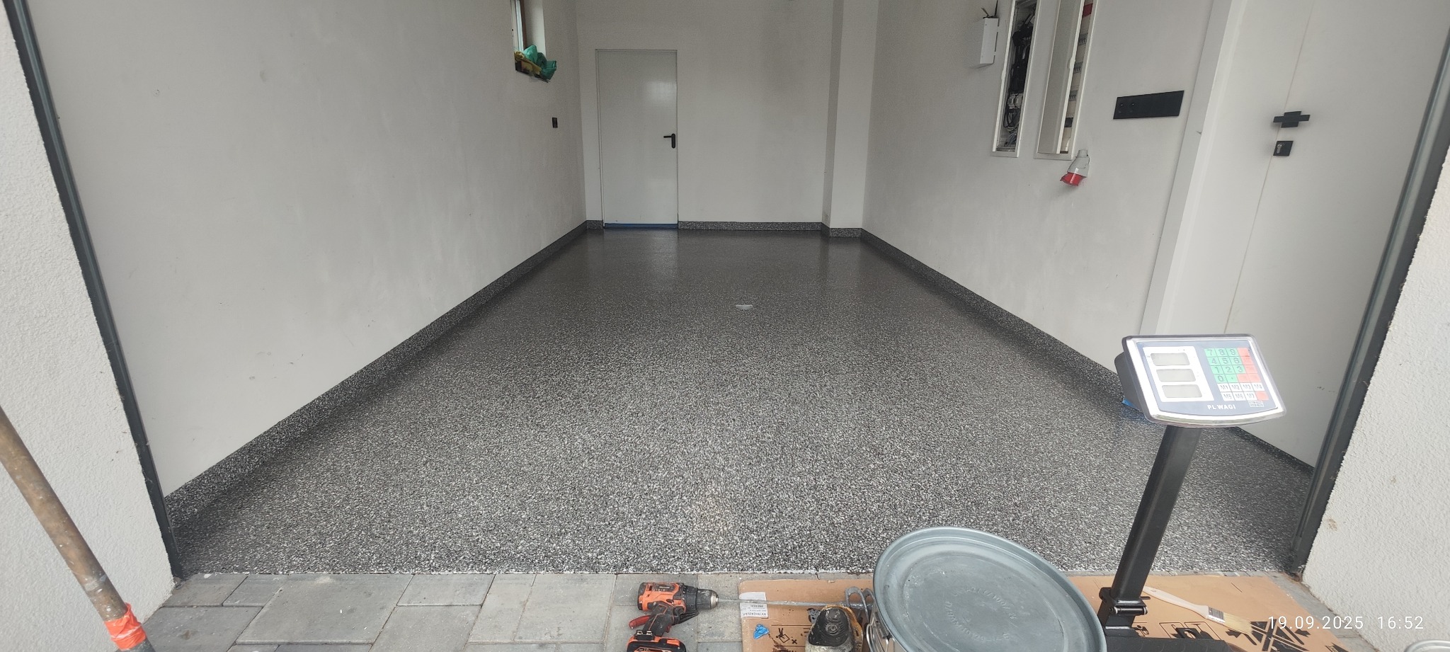 Gładka, szara posadzka żywiczna w garażu, z widocznymi narzędziami pracy: wiertarką, puszką z farbą i wagą. Świeżo wykonana, błyszcząca powierzchnia.