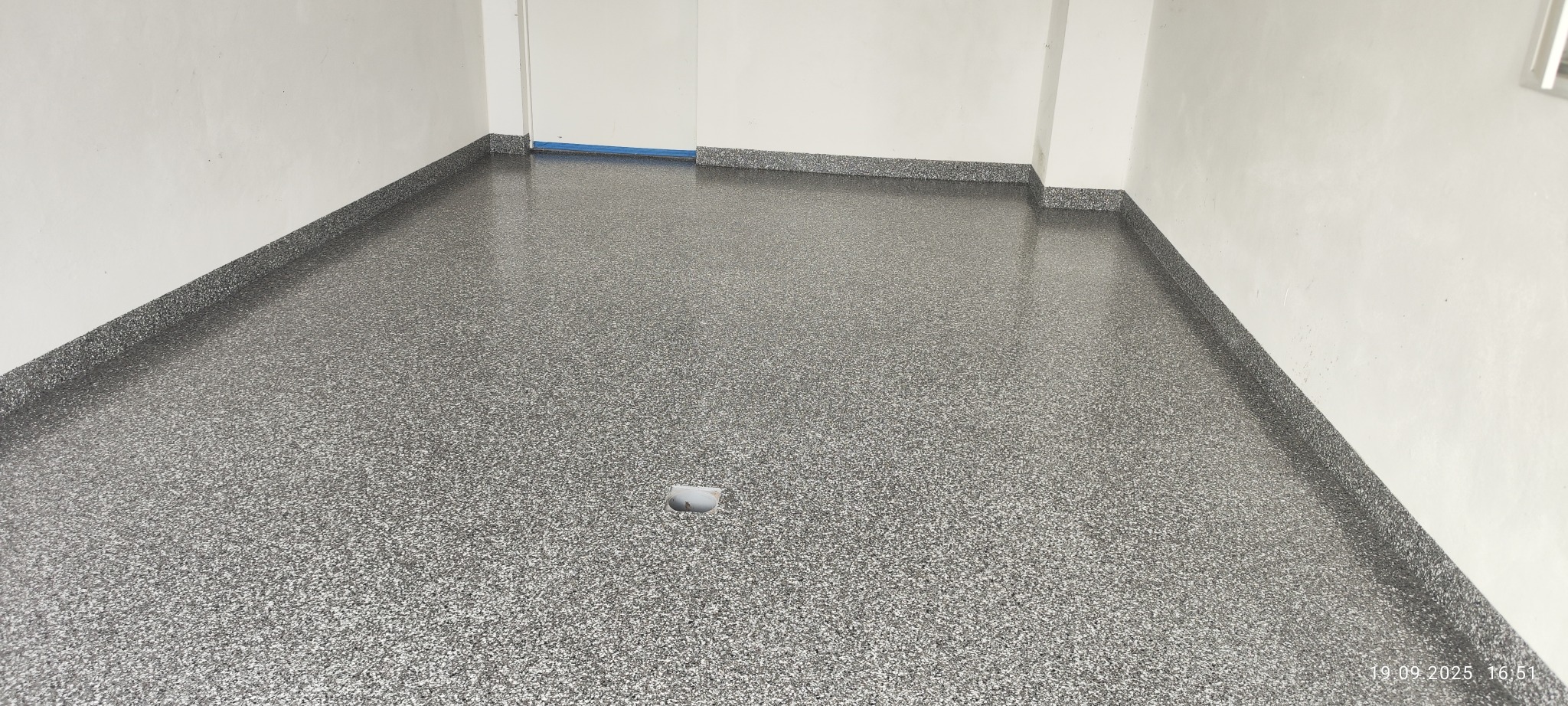 Błyszcząca, szara posadzka z drobnym wzorem w pomieszczeniu z białymi ścianami. Widoczny odpływ podłogowy i fragment niebieskiej taśmy zabezpieczającej.