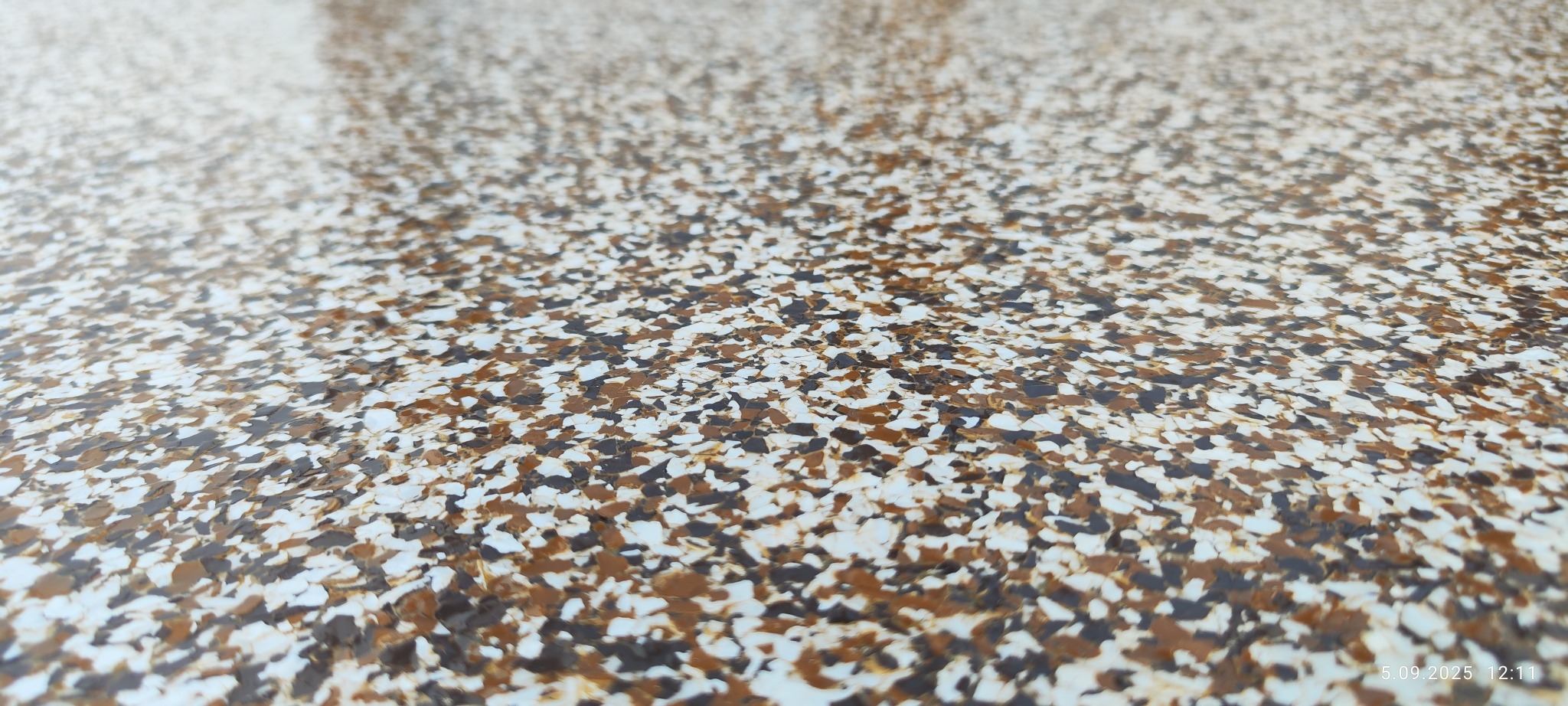 Błyszcząca, gładka powierzchnia z żywicy epoksydowej z dekoracyjnymi płatkami w odcieniach brązu i bieli, tworząca unikalny wzór na podłodze.