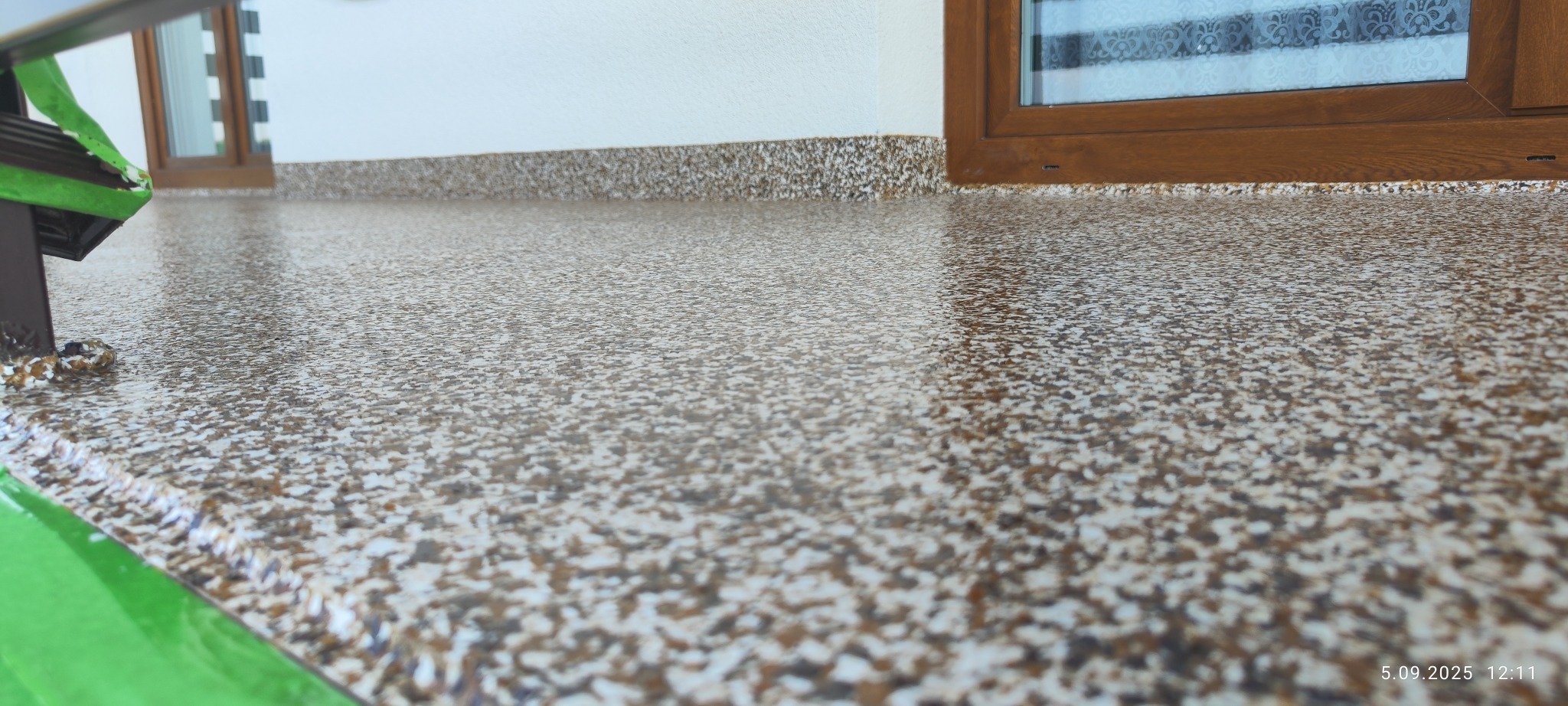 Błyszcząca, brązowo-biała posadzka żywiczna na balkonie, widoczna z niskiego kąta. W tle okno z brązową ramą i biała ściana. Na pierwszym planie fragment balustrady z zielonym zabezpieczeniem.