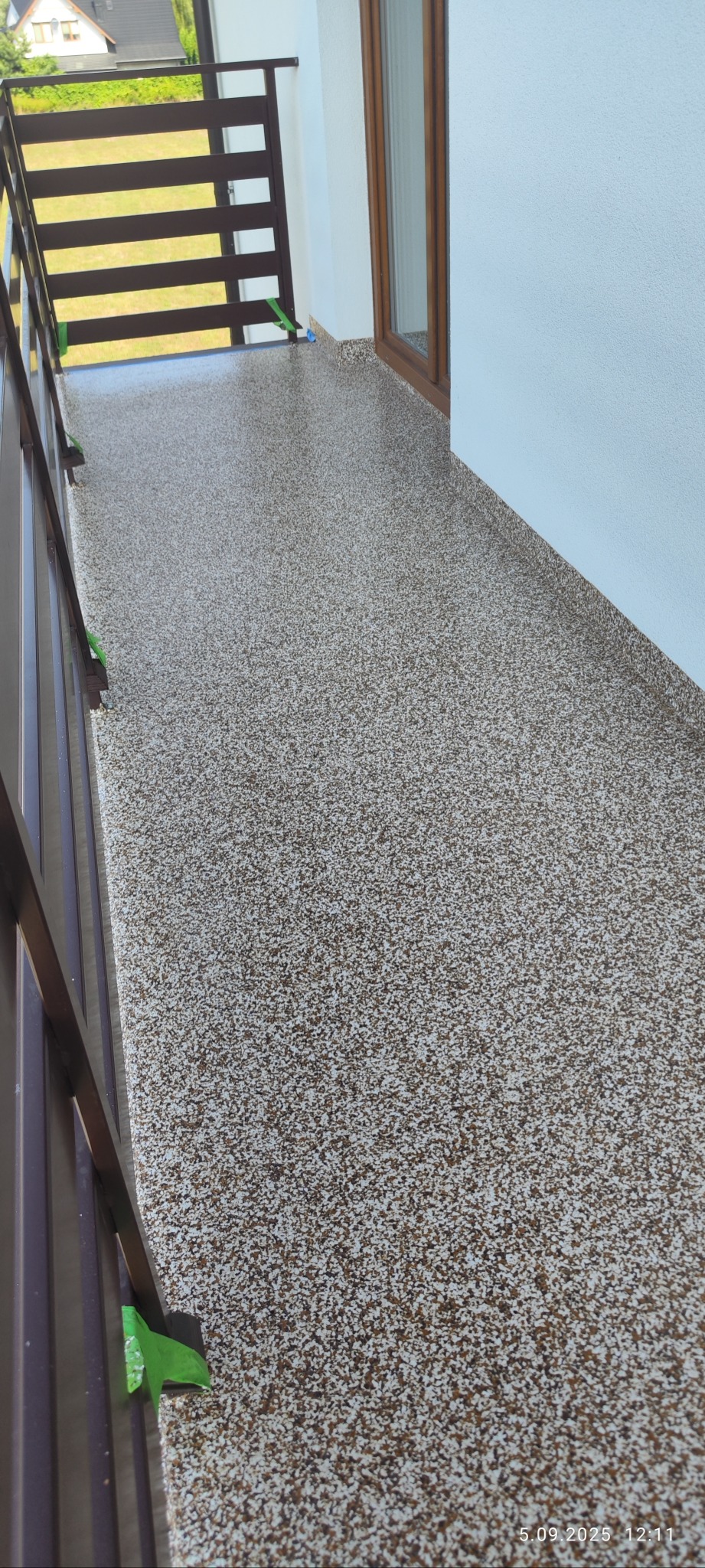 Balkon z beżowo-brązową, wzorzystą posadzką. Widoczne brązowe barierki i fragment okna. Na barierce zielona taśma zabezpieczająca.