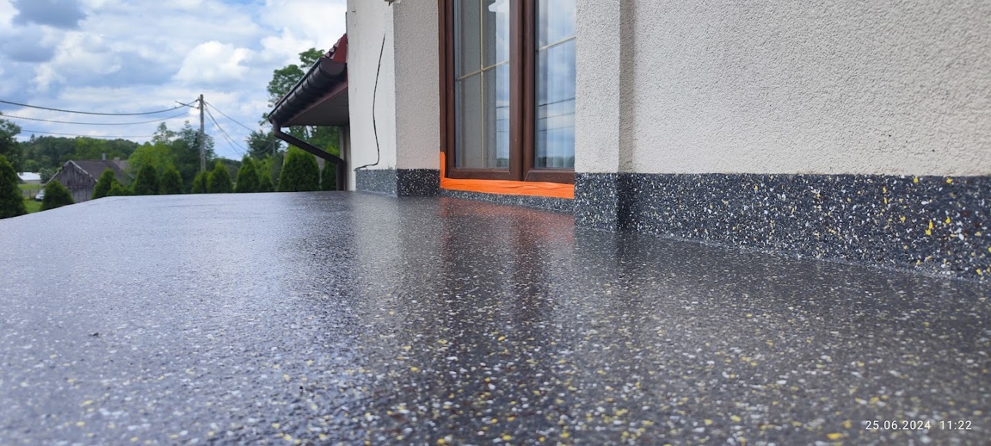 Błyszcząca, szara posadzka z drobnymi, kolorowymi elementami na balkonie. Widoczne okno z brązową ramą i fragment elewacji budynku. Tło stanowią drzewa i błękitne niebo.