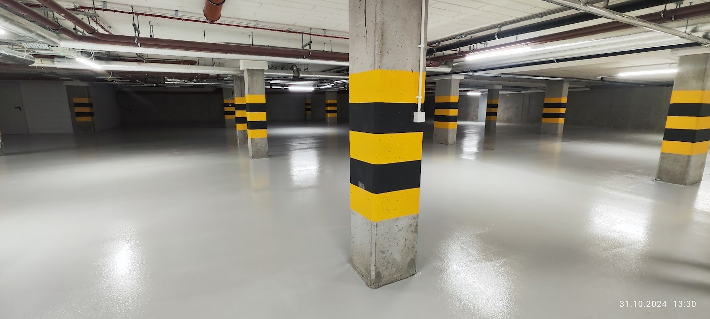 Jasnoszara, błyszcząca posadzka w garażu podziemnym. Betonowe słupy pomalowane w czarno-żółte pasy. Widoczne rury pod sufitem i oświetlenie. Gładka powierzchnia.