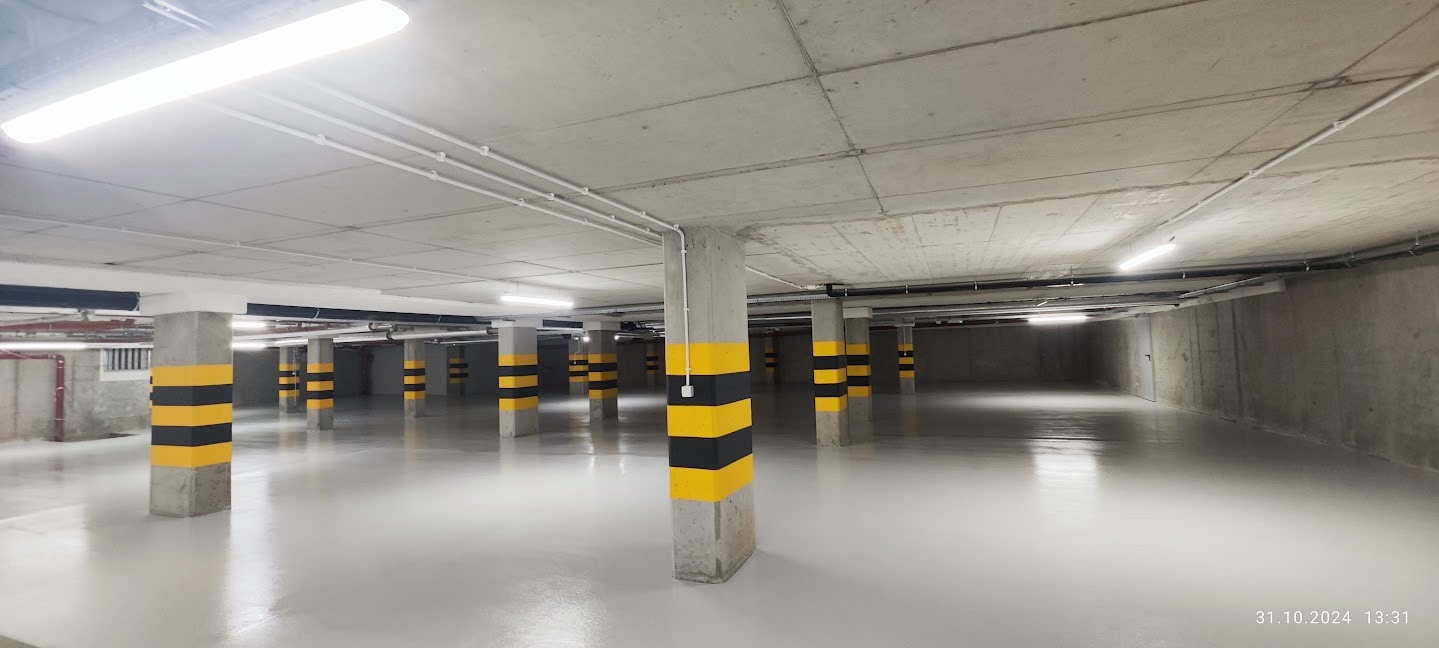 Jasnoszara, gładka posadzka w garażu podziemnym z betonowymi kolumnami w czarno-żółte pasy. Widoczne oświetlenie sufitowe i instalacje. Perspektywa z głębi garażu.