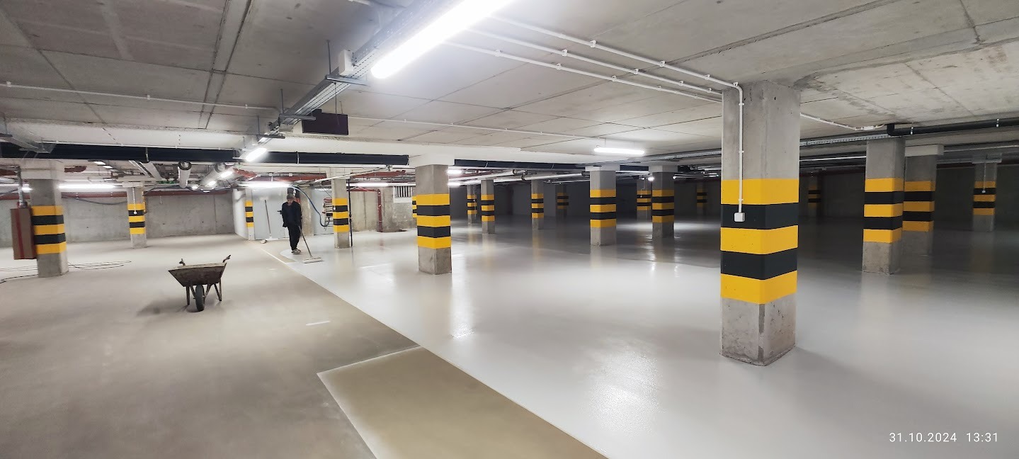Jasna posadzka w garażu podziemnym z kolumnami w pasy. Pracownik rozprowadza żywicę. W tle wózek budowlany. Widoczne instalacje pod sufitem.