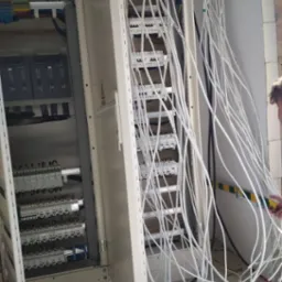 Otwarta szafa elektryczna z licznymi białymi kablami, widoczne zabezpieczenia i złącza. Część kabli wychodzi poza obrys szafy, ściana w tle.