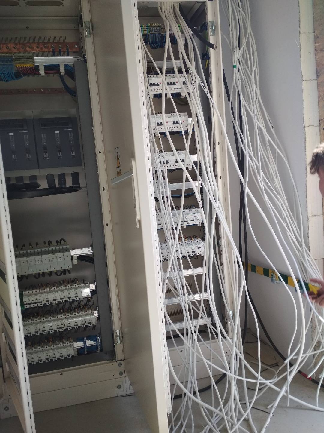 Otwarta szafa elektryczna z licznymi białymi kablami, widoczne zabezpieczenia i złącza. Część kabli wychodzi poza obrys szafy, ściana w tle.