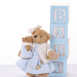 Anna Stankiewicz - Pluszowy miś w błękitnej sukience trzyma mniejszego misia, obok wieża z drewnianych klocków z literami 'BABY'. Jasne tło, delikatna faktura podłoża.