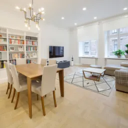 Jasne wnętrze salonu z jadalnią w Krakowie. Parkiet w jodełkę, st&oacute;ł z krzesłami, regał z książkami, sofa i dywan z geometrycznym wzorem. Nowoczesny żyrandol.