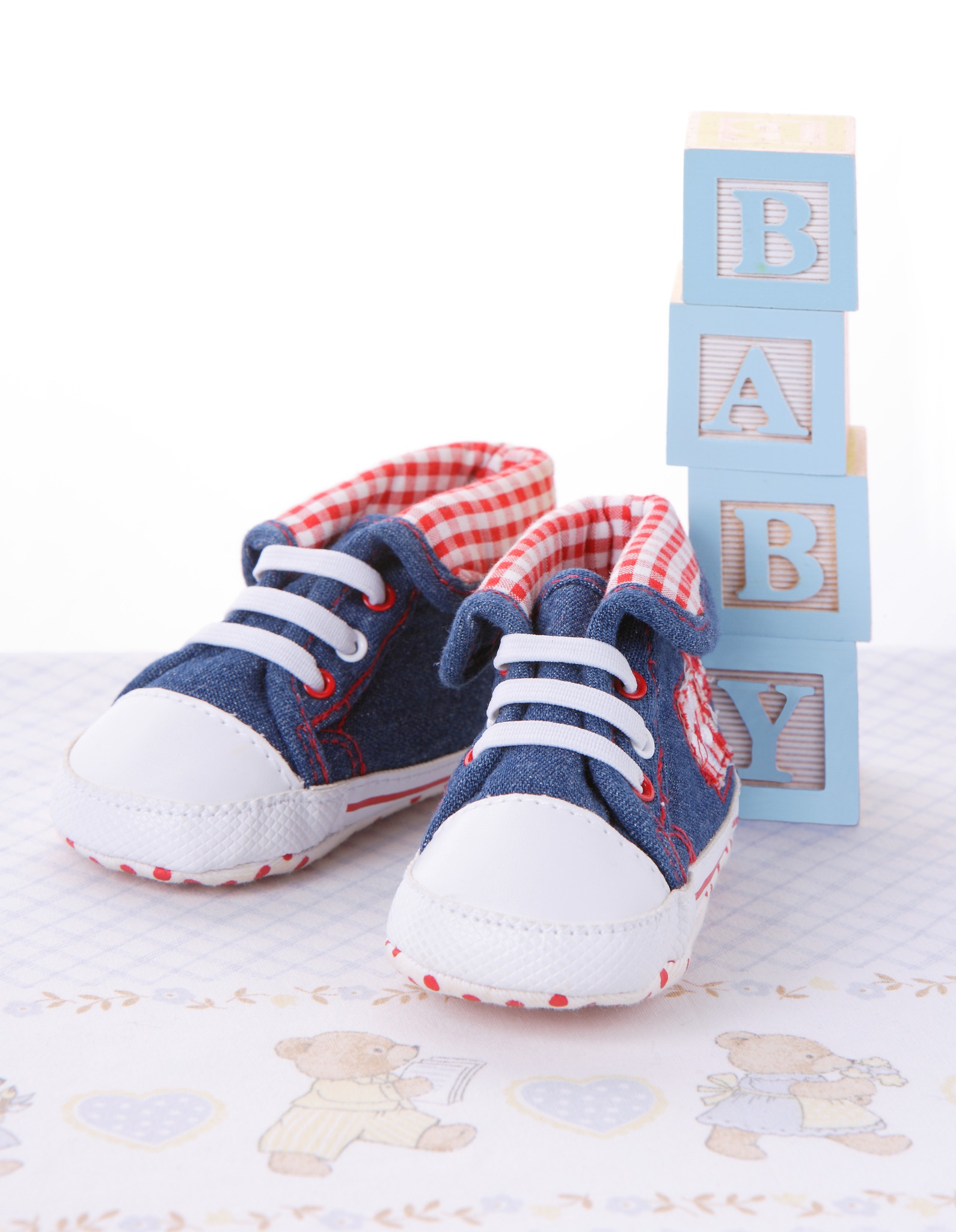 Dziecięce, dżinsowe buciki z białą gumą i czerwoną podszewką w kratę, stojące obok wieży z drewnianych, błękitnych klocków z literami 'BABY' na tle koca w misie.