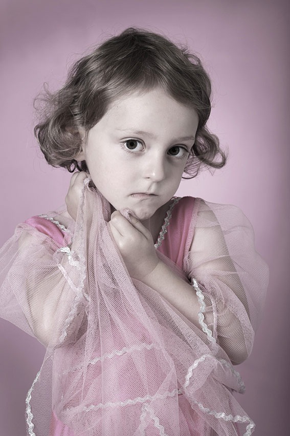 Portret dziewczynki w różowej sukience z tiulem na tle pudrowego różu. Ujęcie z bliska, skupione na wyrazie twarzy i delikatnych detalach stroju.