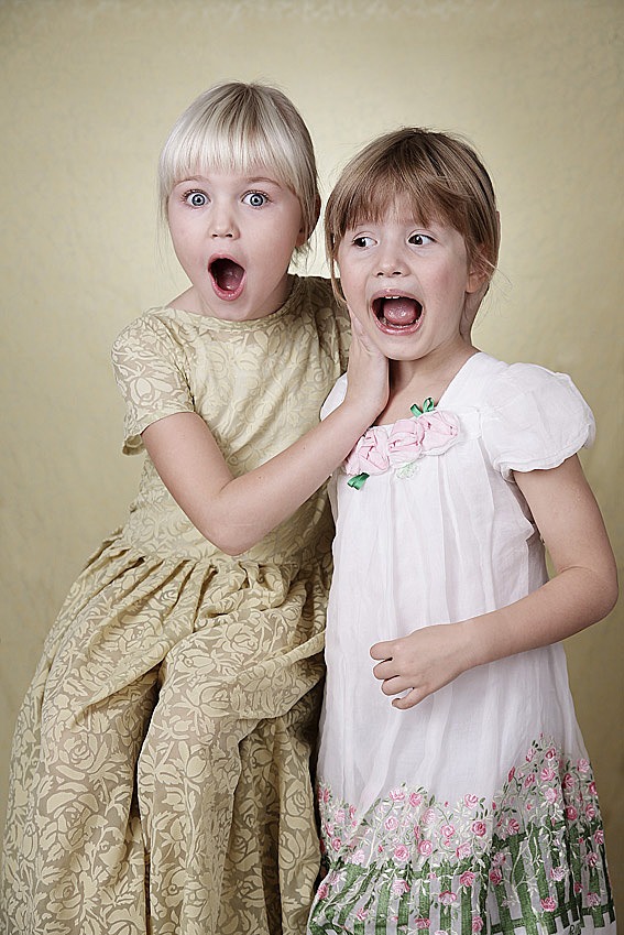 Dwie dziewczynki w sukienkach, z szeroko otwartymi ustami, wyrażające zaskoczenie na tle jednolitego, jasnego tła. Blondynka w kwiecistej sukni dotyka policzka brunetki.