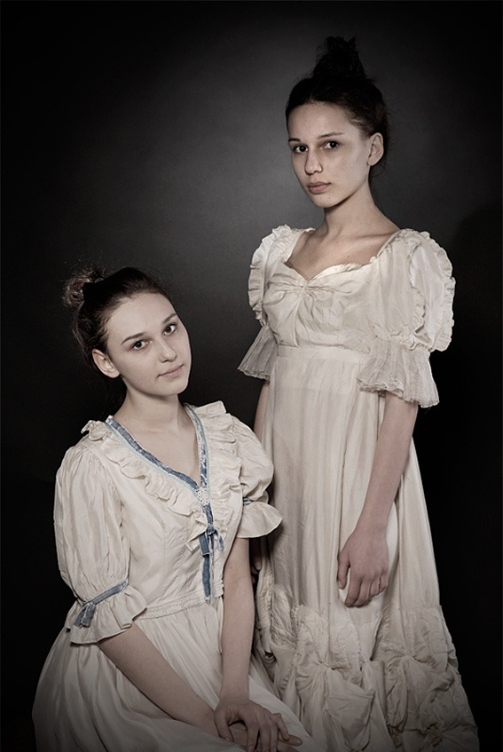 Portret dwóch młodych kobiet w stylizowanych, jasnych sukniach na ciemnym tle. Ujęcie studyjne, skupiające się na wyrazie twarzy i detalach ubioru.