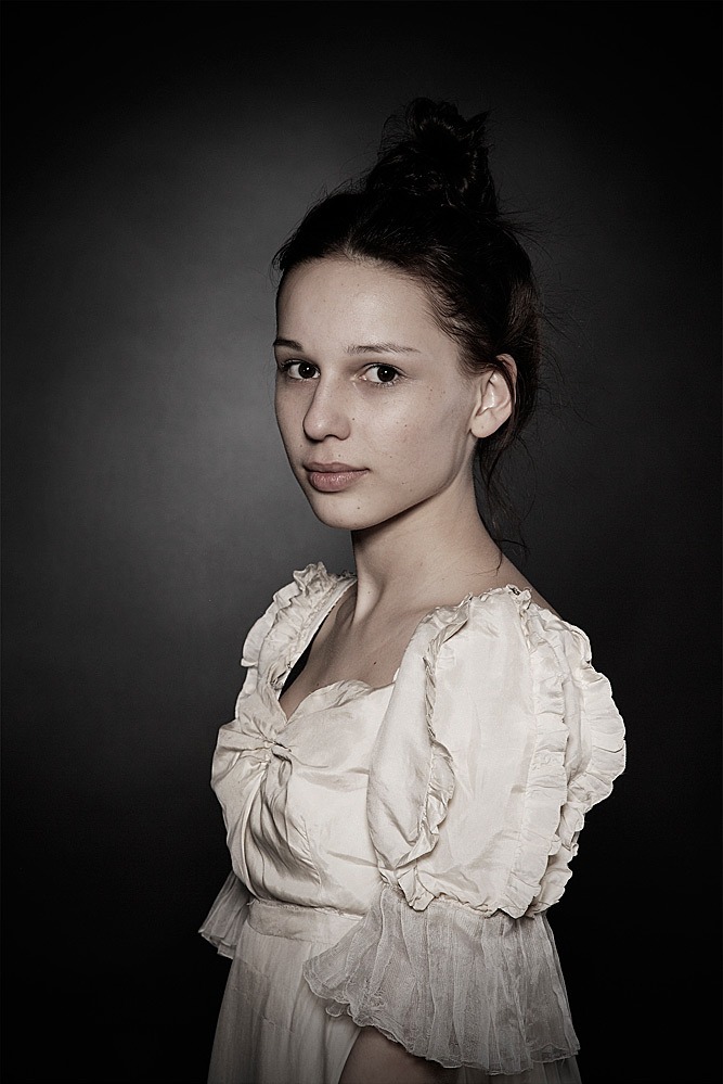 Portret młodej kobiety w jasnej sukience z bufkami na ciemnym tle. Uczesane włosy, delikatny makijaż, stonowana kolorystyka nadają zdjęciu klasyczny charakter.