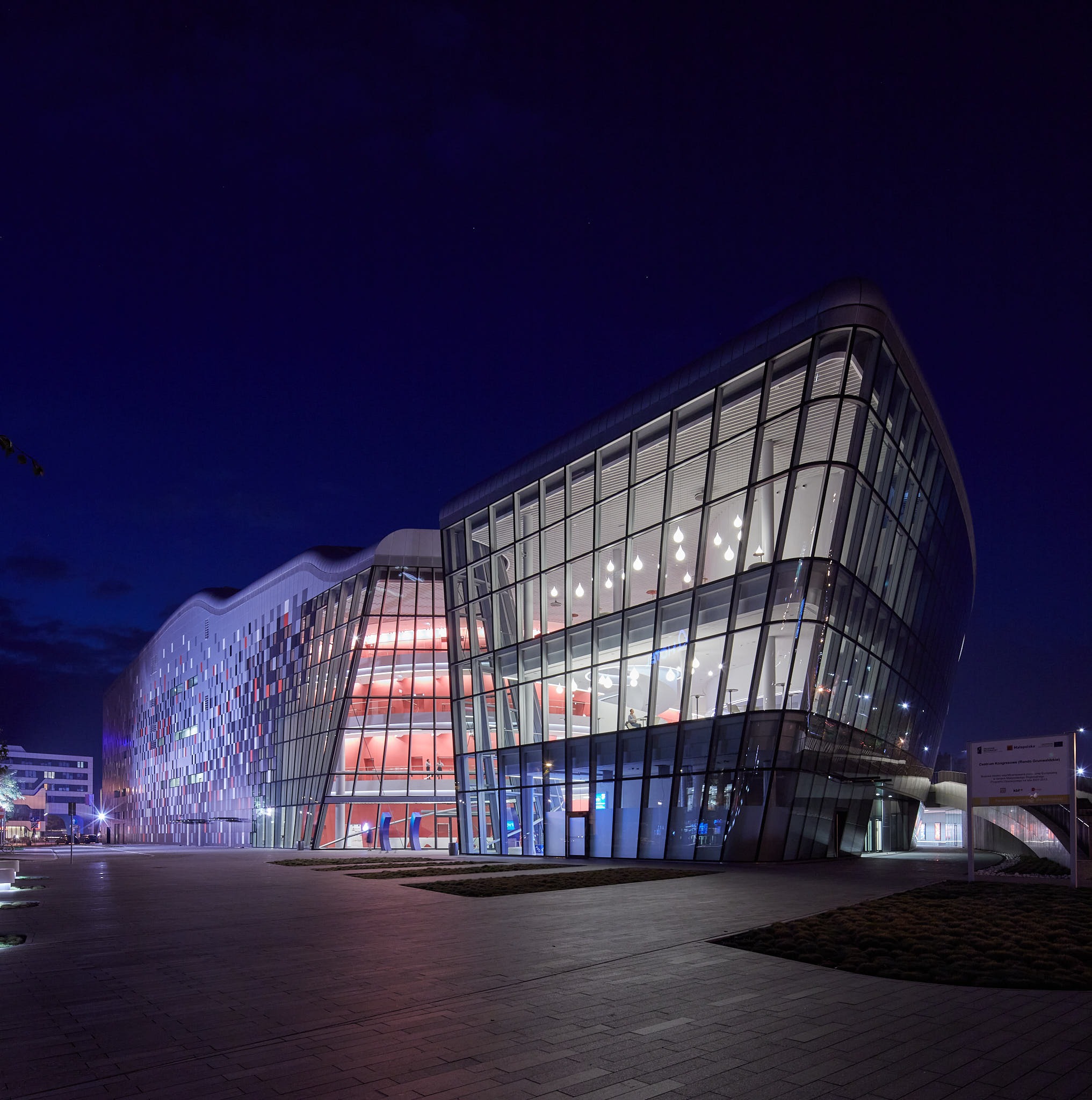 Nowoczesny budynek kongresowy ICE Kraków nocą, z widocznymi oknami i oświetleniem wewnętrznym, na tle ciemnego, granatowego nieba. Unikalna architektura.