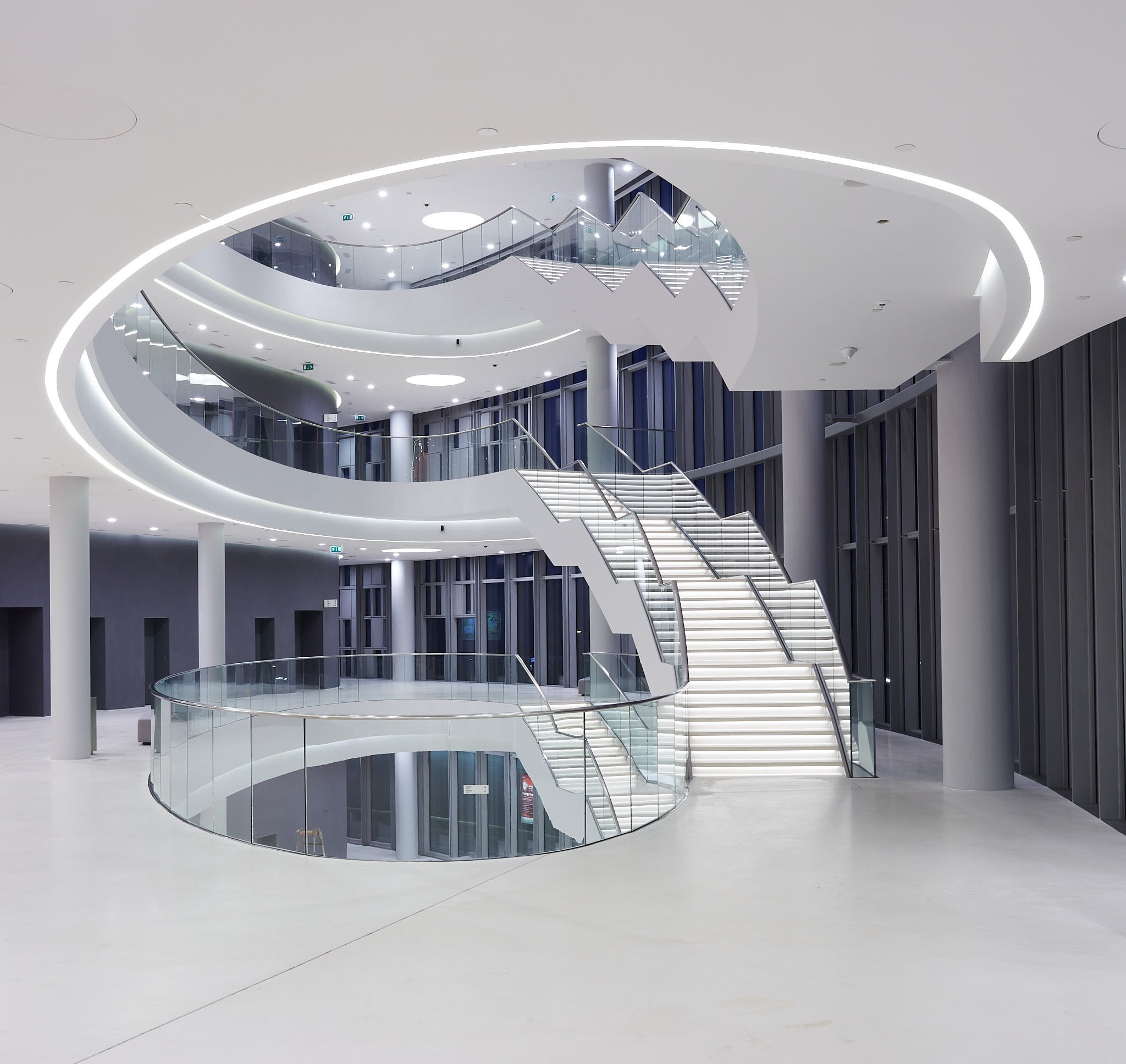 Nowoczesne, jasne wnętrze z owalnymi schodami oświetlonymi ledami, szklane balustrady, kolumny i minimalistyczny design. Widok z dołu na górę.