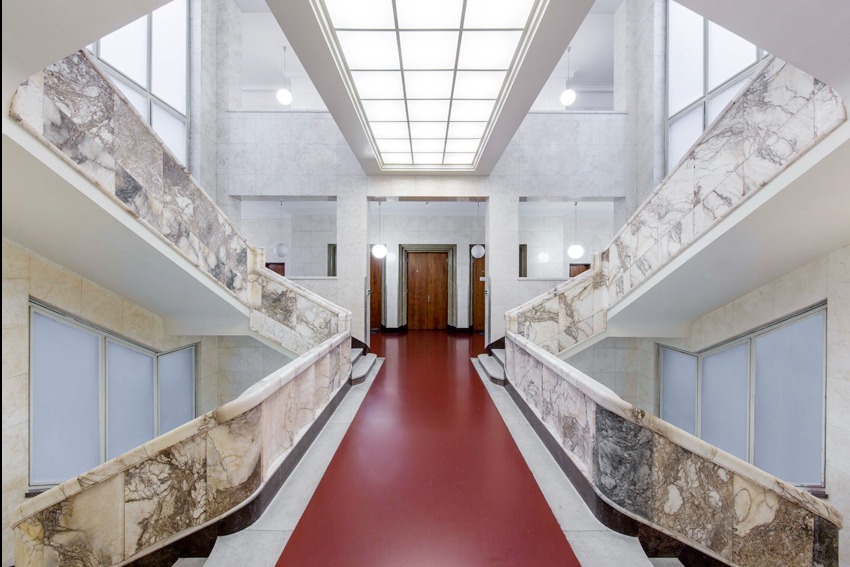 Symetryczne ujęcie marmurowych schodów z czerwoną wykładziną, oświetlonych lampami i plafonem, w jasnym, modernistycznym wnętrzu budynku.