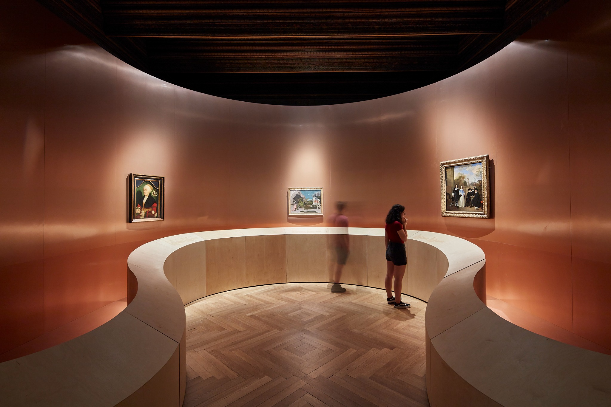 Wnętrze galerii sztuki z drewnianą podłogą w jodełkę. Na ścianach wiszą obrazy w ozdobnych ramach. Dwie osoby oglądają ekspozycję.