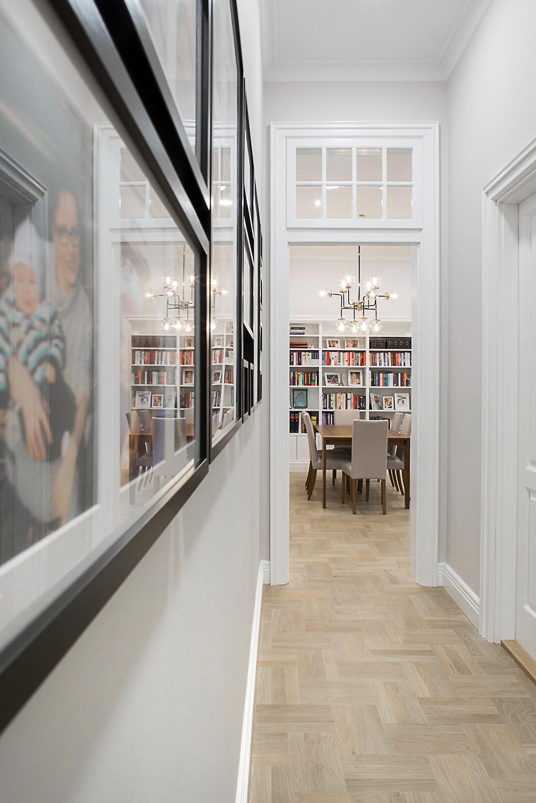 Wnętrze hallu z parkietem w jodełkę, ściany zdobią ramy ze zdjęciami. W głębi widoczny pokój z biblioteczką i stołem jadalnym pod nowoczesnym żyrandolem.