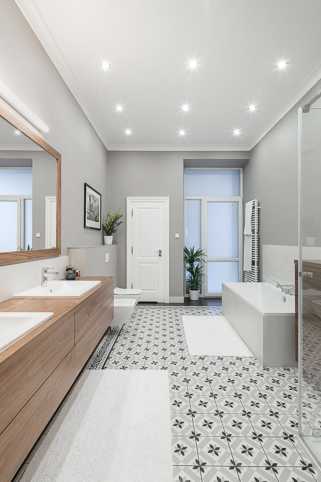 Nowoczesna łazienka w szarościach z drewnianymi akcentami. Uwagę przyciągają wzorzyste płytki podłogowe i duże lustro nad podwójną umywalką. Jasne, przestronne wnętrze.