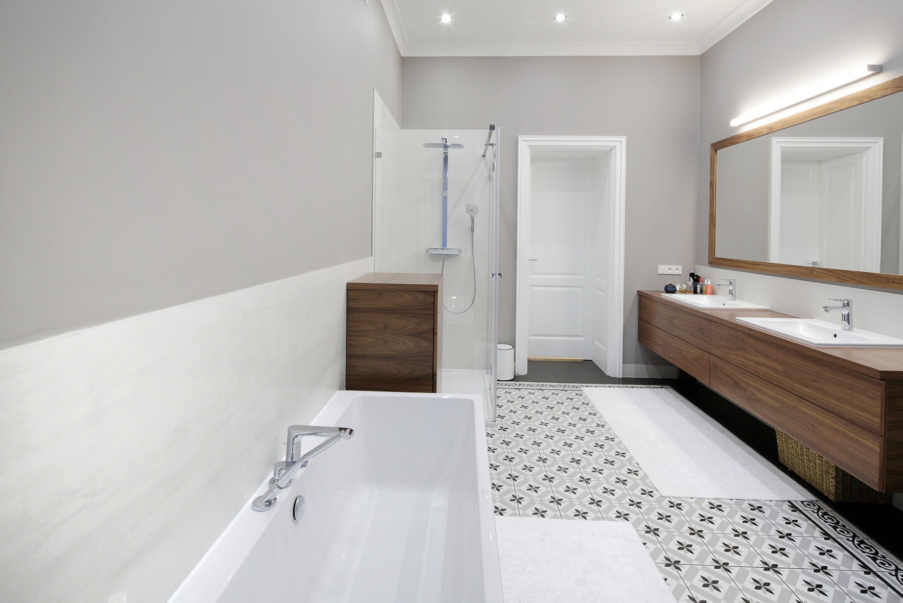 Nowoczesna łazienka z drewnianymi akcentami: szafka pod umywalką i obudowa prysznica w odcieniach brązu, biała wanna oraz wzorzysta podłoga.