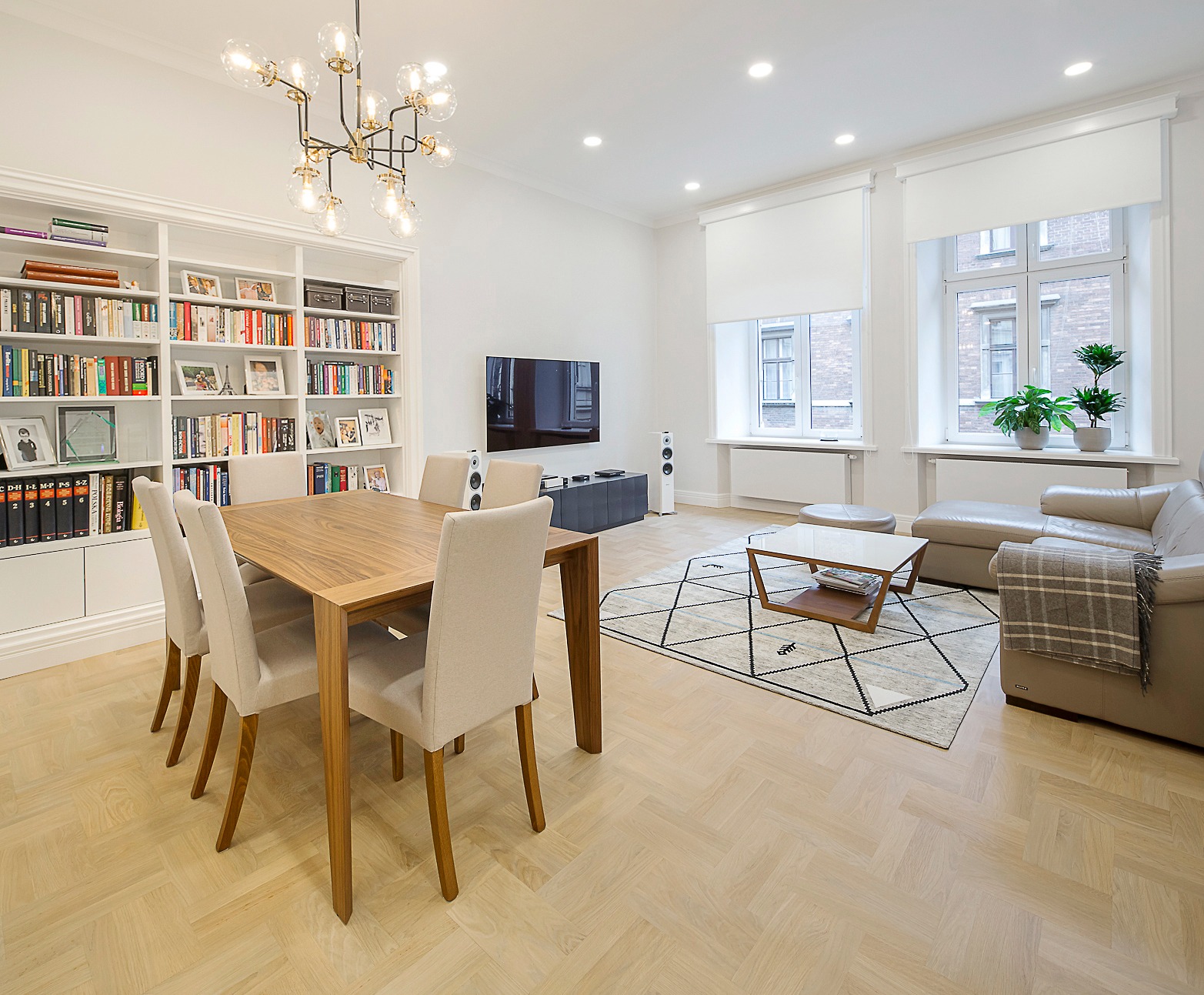 Jasne wnętrze salonu z jadalnią w Krakowie. Parkiet w jodełkę, stół z krzesłami, regał z książkami, sofa i dywan z geometrycznym wzorem. Nowoczesny żyrandol.