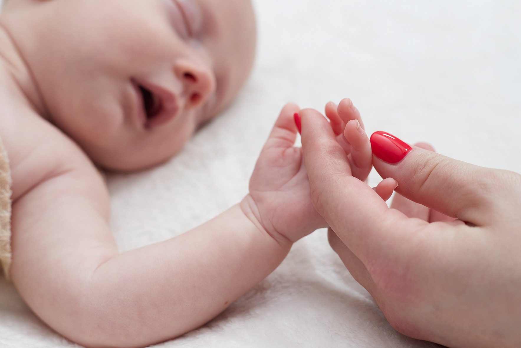 Śpiące niemowlę trzyma za rękę dorosłą osobę z czerwonymi paznokciami na tle białego koca. Delikatne ujęcie, bliski kadr, ciepłe światło.