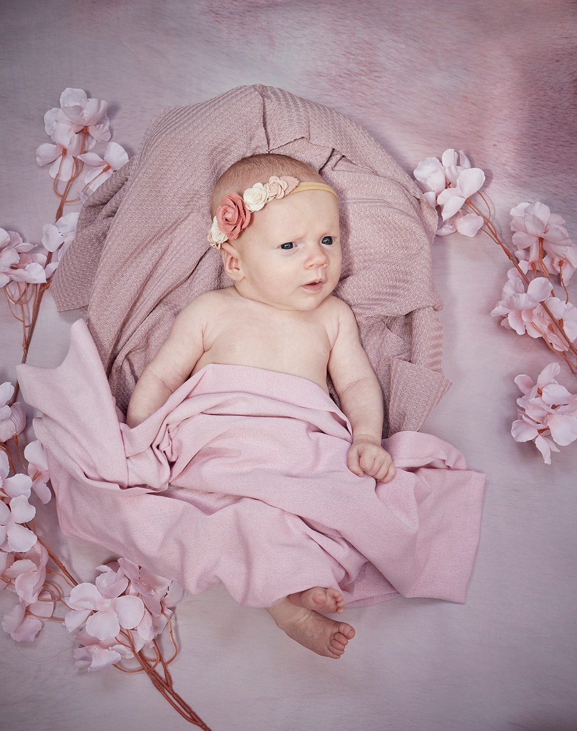 Niemowlę z opaską kwiatową, otulone różowym kocykiem, otoczone gałązkami kwitnącej wiśni na tle w odcieniach różu. Ujęcie z góry, miękkie światło.