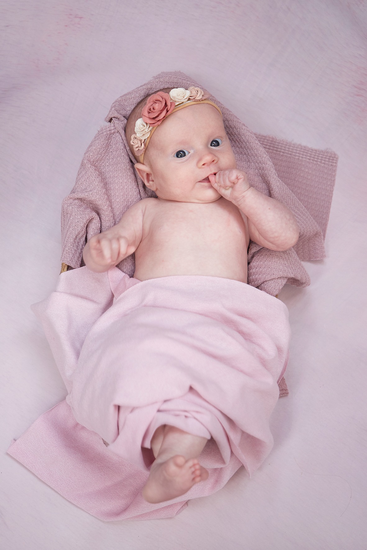 Niemowlę z opaską z kwiatami, owinięte w różowy kocyk, leży na białym tle. Dziecko ssie palec, patrzy w górę, delikatne oświetlenie.